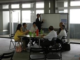 基本設計ワークショップで1人がホワイトボードの前に立ち机を囲んで6名が話し合いをしている写真