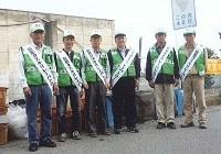 緑のベストを着て襷をかけている6人の男性が並んでいる写真