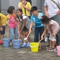 バケツに入った水を柄杓で打ち水をしている子供たちの写真