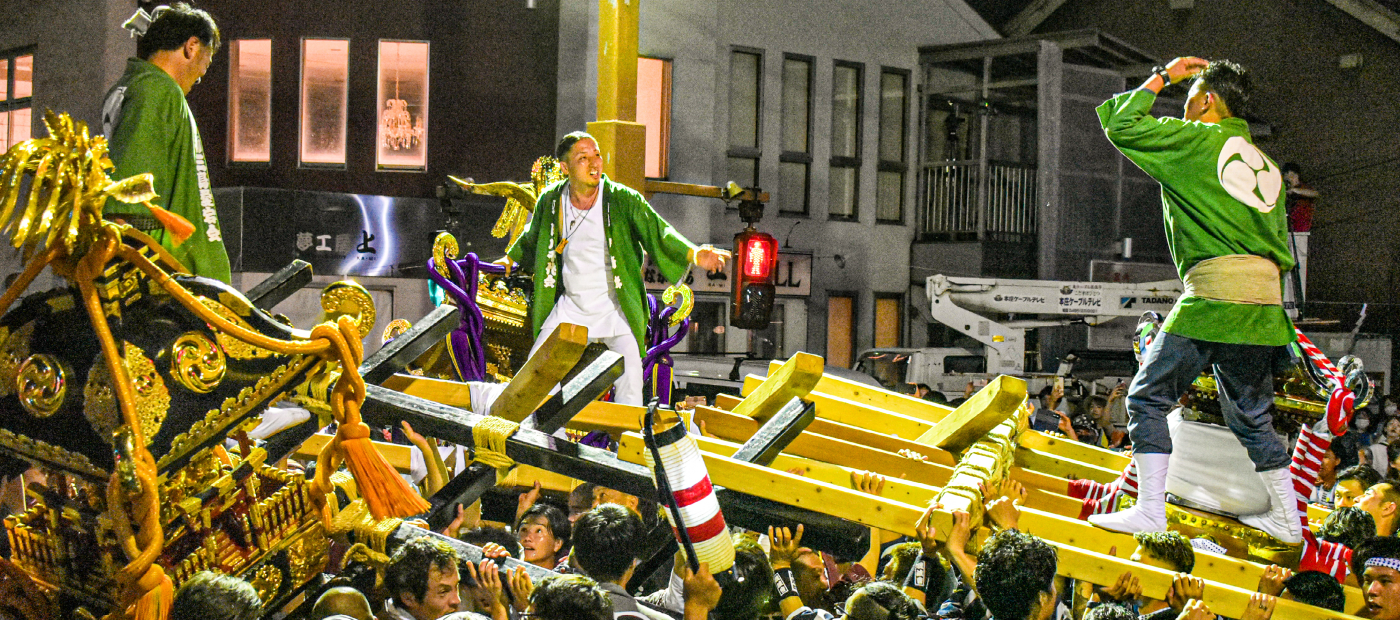 夜の祭りの様子 大勢で神輿を担いでおり、その神輿の上に法被を着た男性3人が立っている