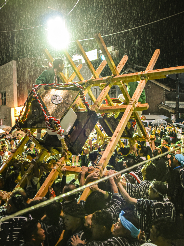 夜の祭りの様子 雨が降る中、大勢で神輿を担いでおりその神輿の上に男性2人が立っている