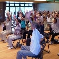 筋力アップ教室で参加者達が椅子に座り左手を挙げ伸ばしている写真