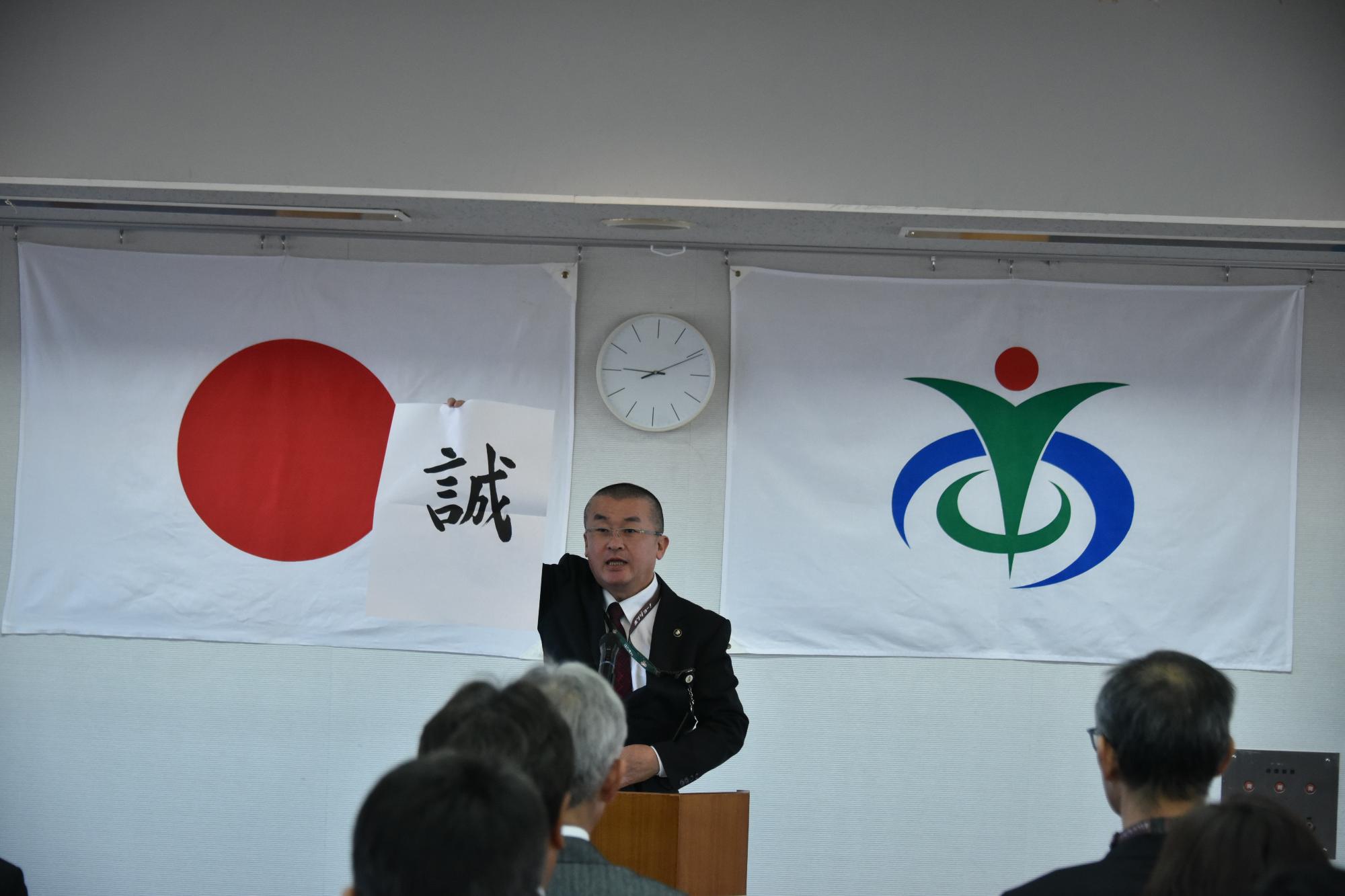 「誠」という文字が書かれた半紙を掲げる本庄市長