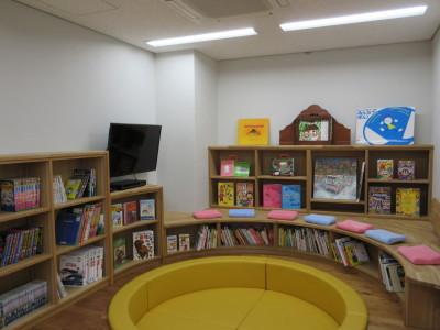 壁際にはテレビや本が並べられ、中央に丸いマットや座布団など座るスペースがある図書室の写真